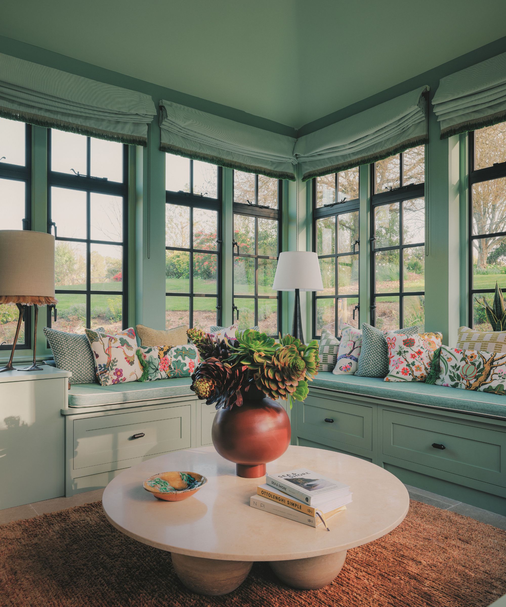 Coin véranda lumineux avec cadres de fenêtres verts, banquette intégrée avec coussins floraux, table basse ronde beige et pièce maîtresse succulente en terre cuite.